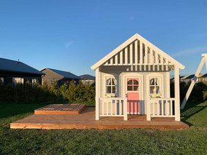a2 disain täispuidust laste mängumaja Rannamaja eestvaade roosa ukse, valgete lillekastide ja  puidust terrassiga